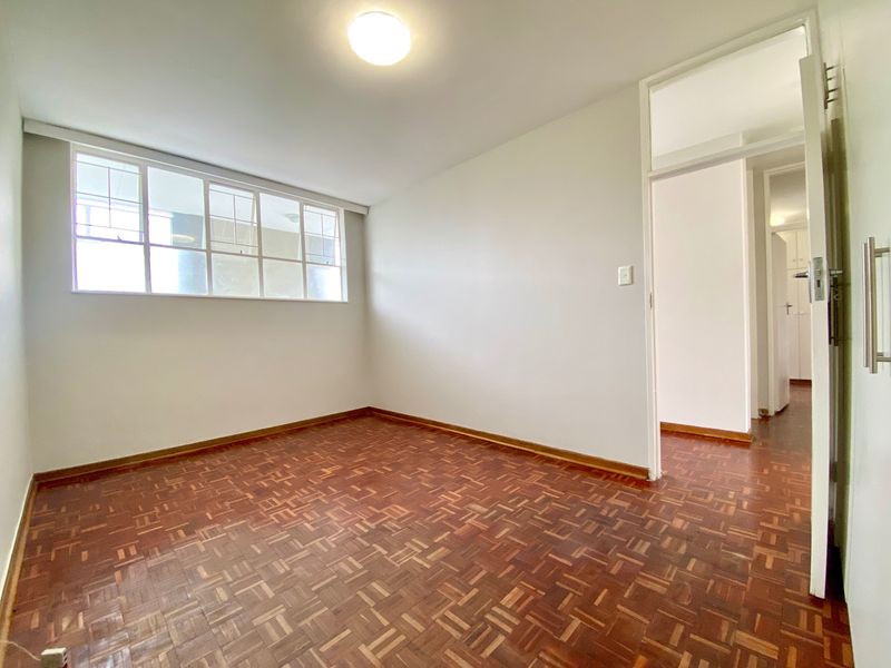 A bright, empty bedroom featuring classic parquet flooring, a large multi-pane window, and neutral walls, offering a clean canvas for personalization.