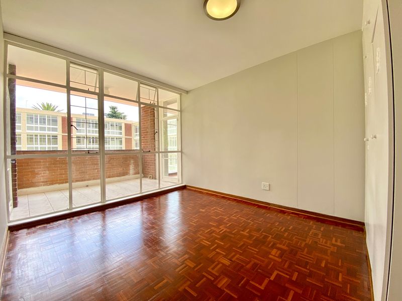 An empty bedroom featuring classic parquet flooring, built-in cupboards, and a large glass wall providing natural light and access to an adjacent balcony.