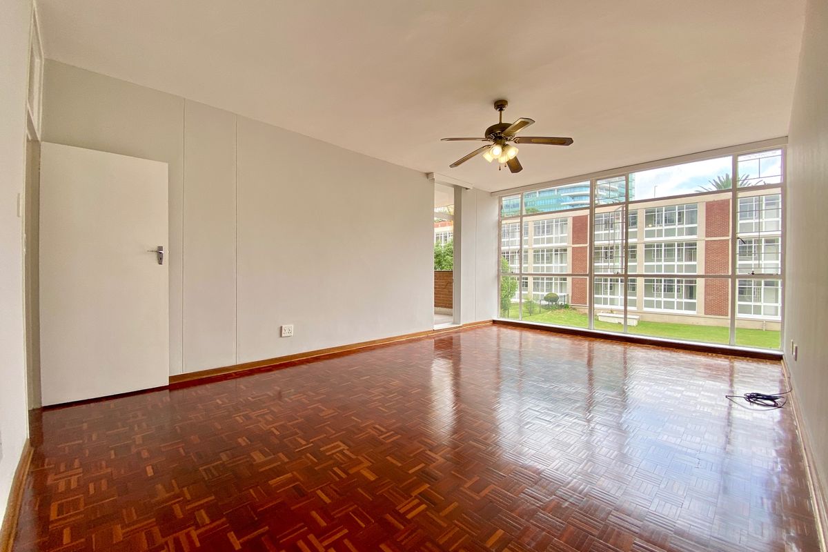 Spacious, empty living room featuring polished parquet flooring, a ceiling fan, and large windows providing ample natural light and views of the complex.