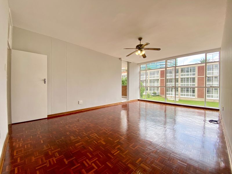 Spacious, empty living room featuring polished parquet flooring, a ceiling fan, and large windows providing ample natural light and views of the complex.
