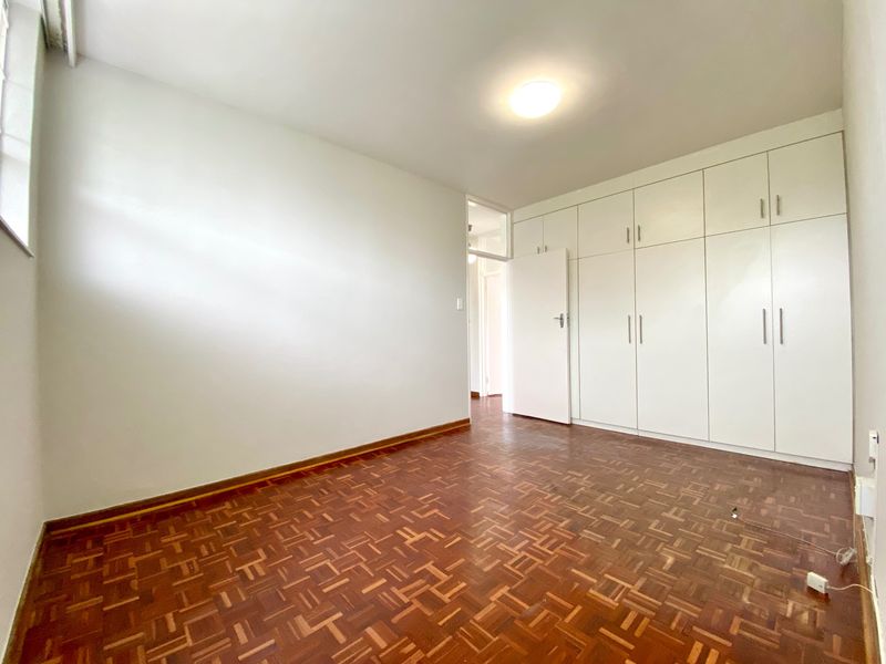 A bright, empty bedroom featuring classic parquet flooring and extensive floor-to-ceiling built-in cupboards, offering ample storage space.
