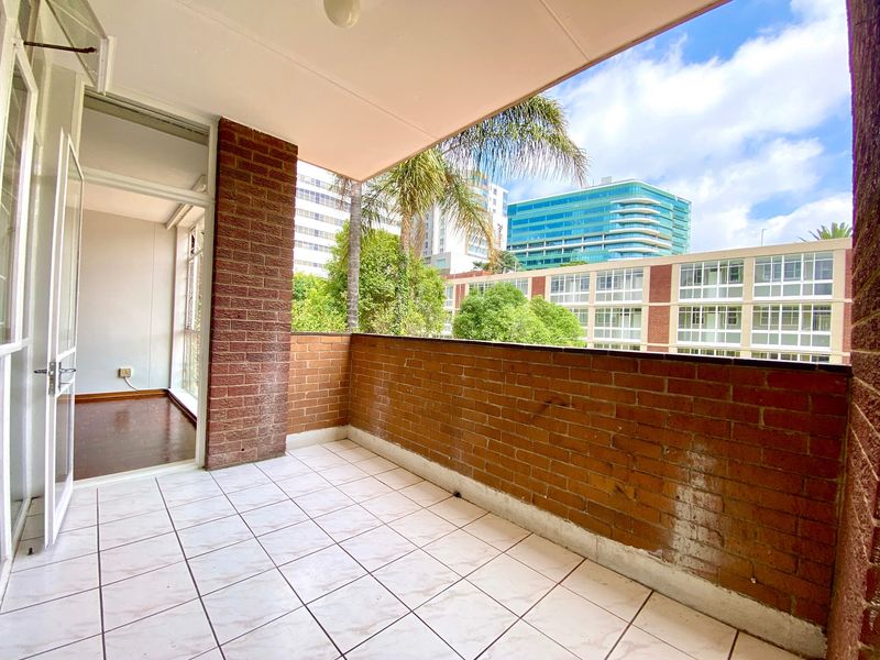 A covered balcony featuring tiled flooring and exposed brick walls, offering an open-air space with views of the surrounding urban environment.