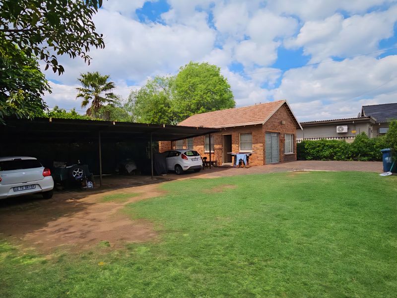 Rear view of the property featuring a brick exterior, a large carport structure, a paved driveway, and a spacious garden area.