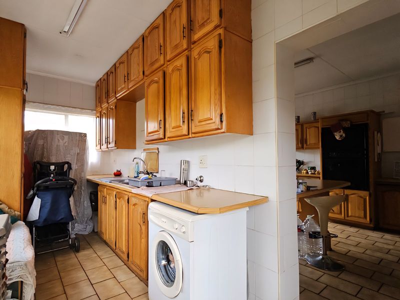 A dated kitchen featuring wooden cabinetry, tiled splashbacks, and tiled flooring, with an adjacent secondary prep or utility area visible through the doorway.