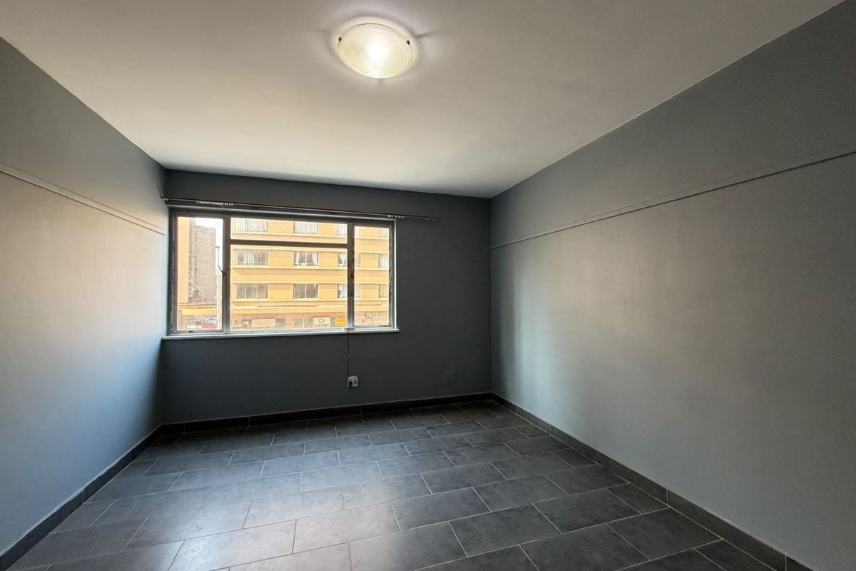 An empty bedroom featuring grey painted walls, dark tiled flooring, and a large window providing natural light.