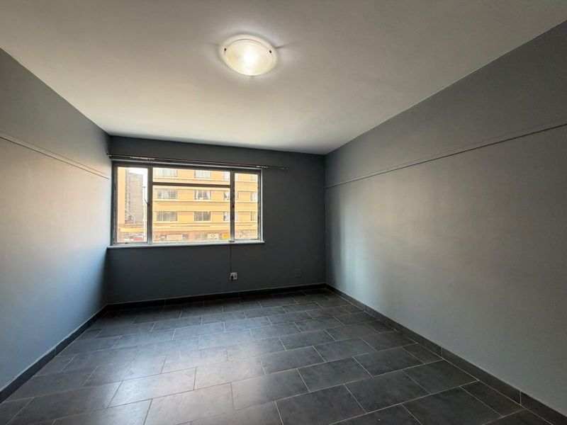 An empty bedroom featuring grey painted walls, dark tiled flooring, and a large window providing natural light.