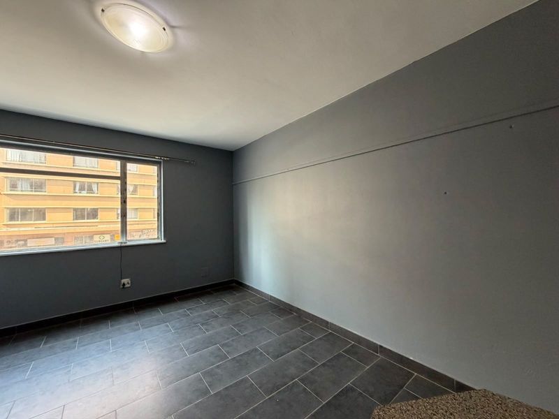 An empty bedroom featuring grey painted walls, dark tiled flooring, and a large window providing natural light.