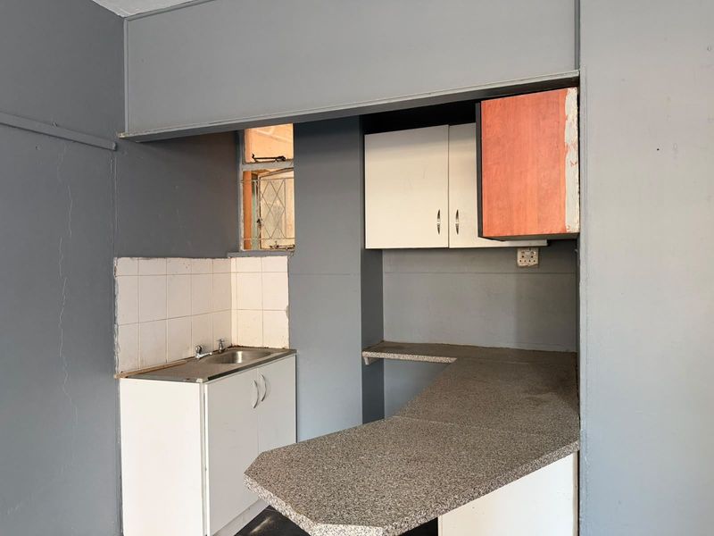 A compact, empty kitchen area featuring built-in cabinetry, a laminate countertop, and a stainless steel sink, requiring cosmetic updates and repairs.