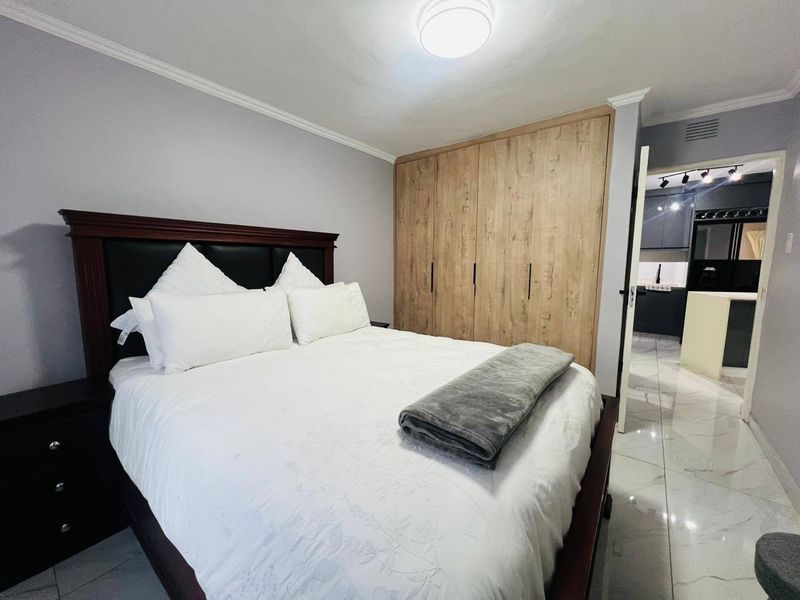 A well-maintained bedroom featuring built-in wooden wardrobes, tiled flooring, and crown molding, with a view into the adjacent kitchen area.
