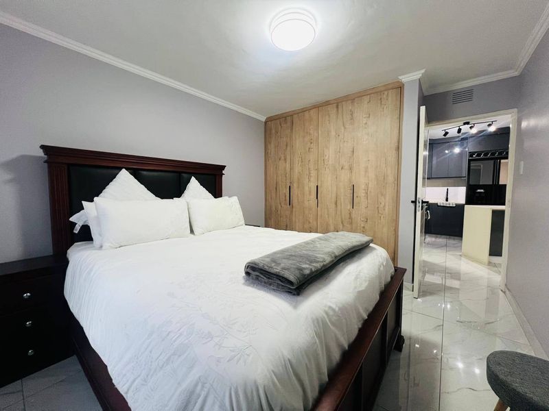 A well-maintained bedroom featuring built-in wooden cupboards, polished tiled flooring, and neutral wall finishes, with an adjacent view into a modern kitchen area.