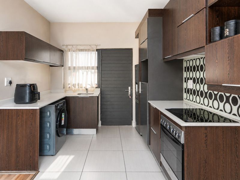 Modern kitchen featuring dark wood cabinetry, a patterned backsplash, integrated electric hob and oven, and light-colored tiled flooring.