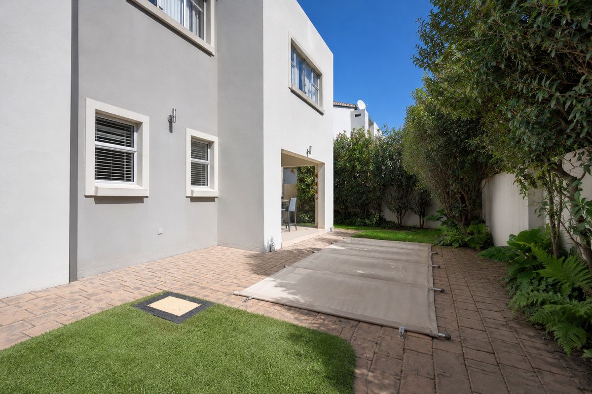 Rear exterior view featuring a paved patio area, covered swimming pool, artificial lawn, and established garden borders.