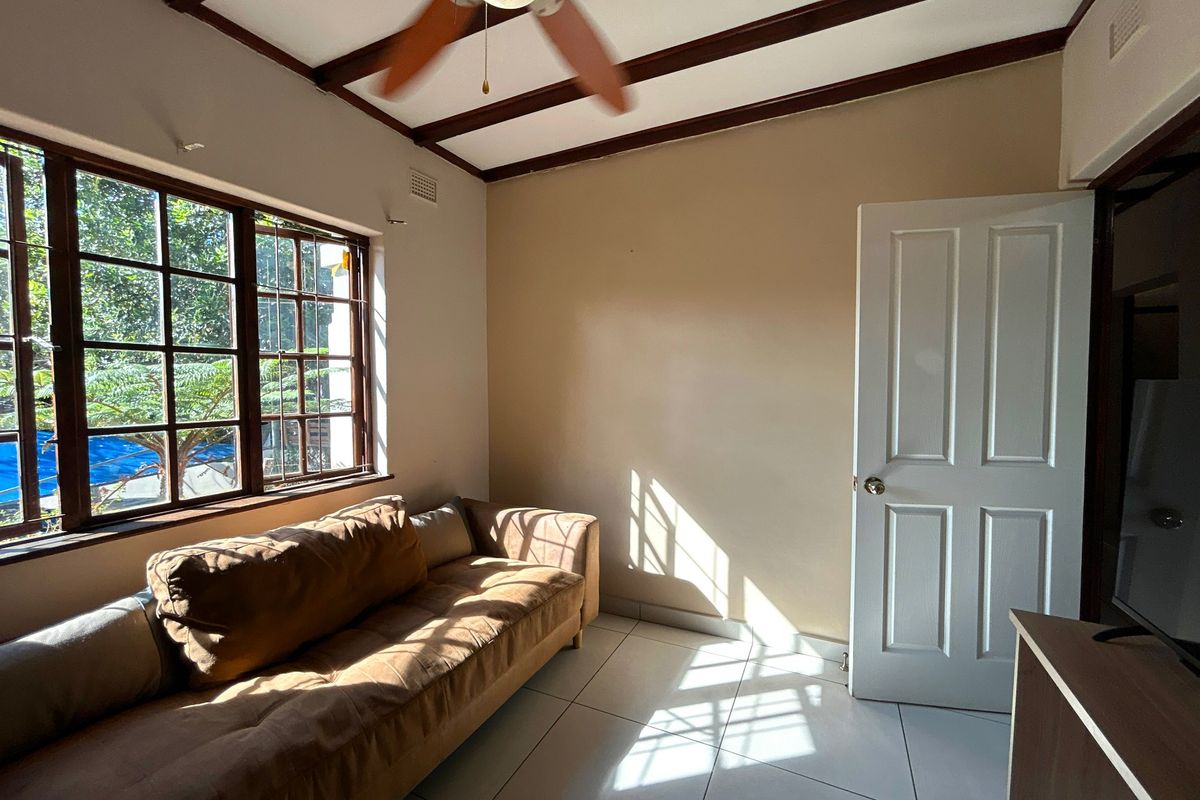 A living room featuring exposed timber ceiling beams, a ceiling fan, tiled flooring, and large multi-pane windows allowing for natural light.