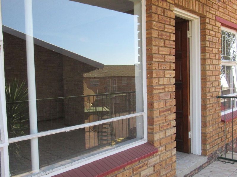 A balcony area featuring face brick exterior walls, tiled flooring, and a metal balustrade, providing access to the interior through a timber door.