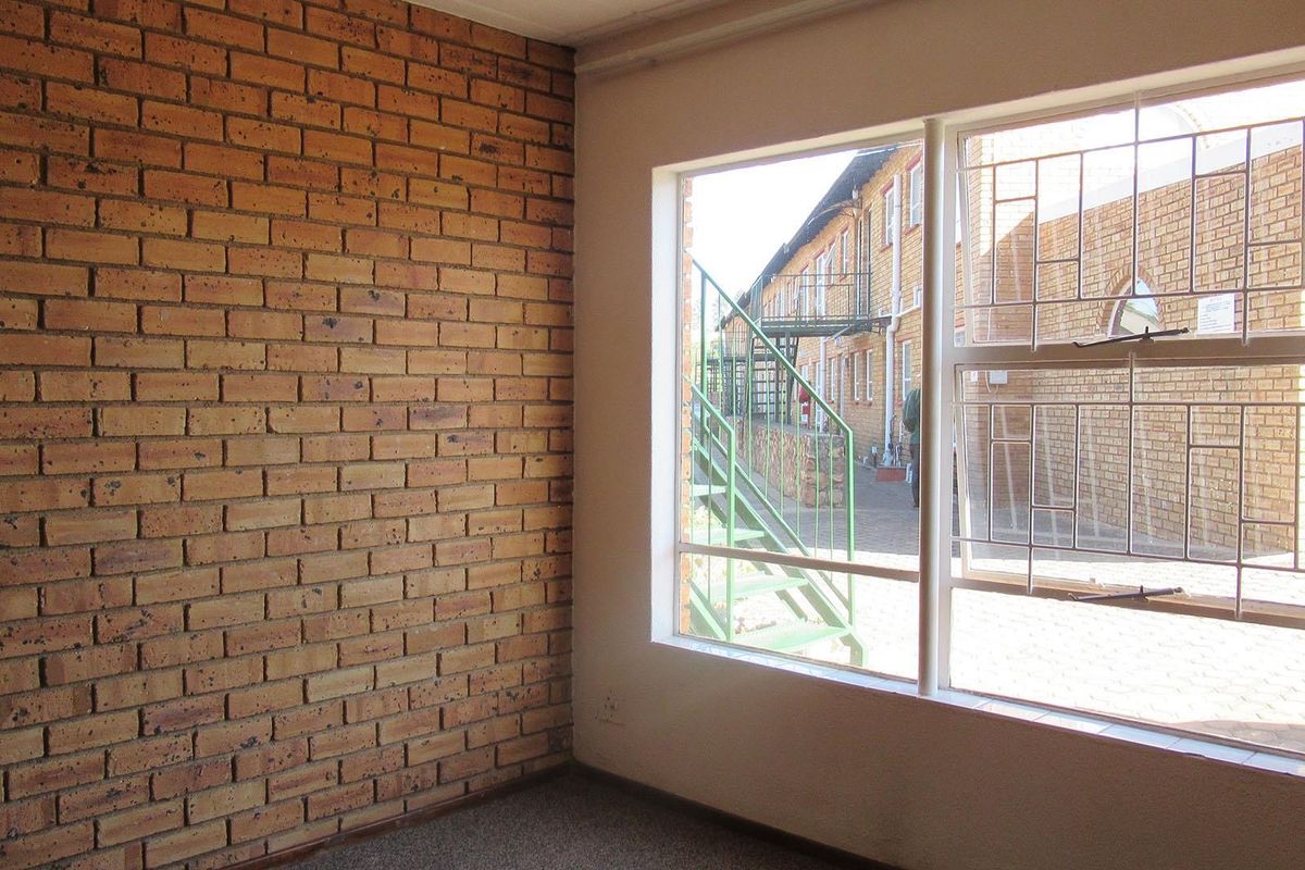 An empty bedroom featuring an exposed brick wall and a large window with security bars, offering a view of the exterior townhouse complex.