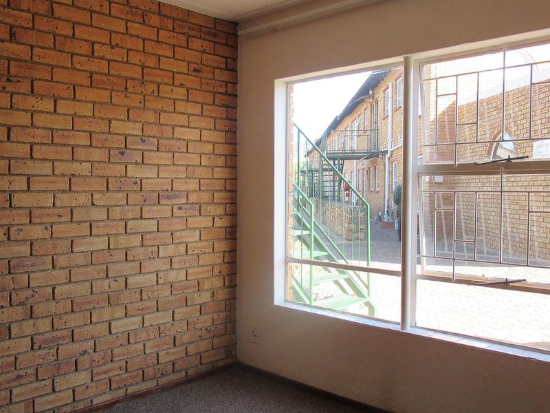 An empty bedroom featuring an exposed brick wall and a large window with security bars, offering a view of the exterior townhouse complex.