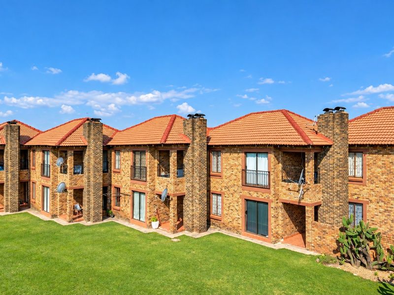 A rear view of a multi-unit apartment complex featuring face brick construction, tiled roofing, and private balconies overlooking a shared lawn area.
