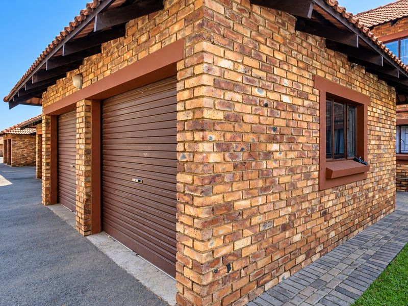 A well-maintained face brick garage block featuring roller shutter doors, tiled roofing, and paved driveway access within a residential apartment complex.