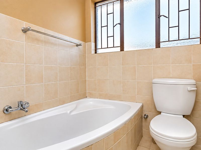A functional bathroom featuring a built-in bathtub, toilet, and neutral wall tiling with a window providing natural light.