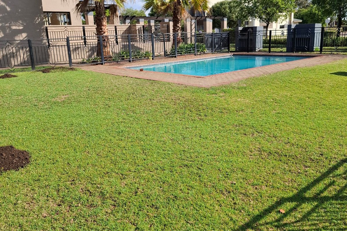 A well-maintained residential backyard featuring a swimming pool with brick coping, surrounded by a manicured lawn and secure metal perimeter fencing.