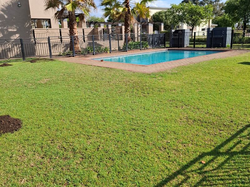 A well-maintained residential backyard featuring a swimming pool with brick coping, surrounded by a manicured lawn and secure metal perimeter fencing.