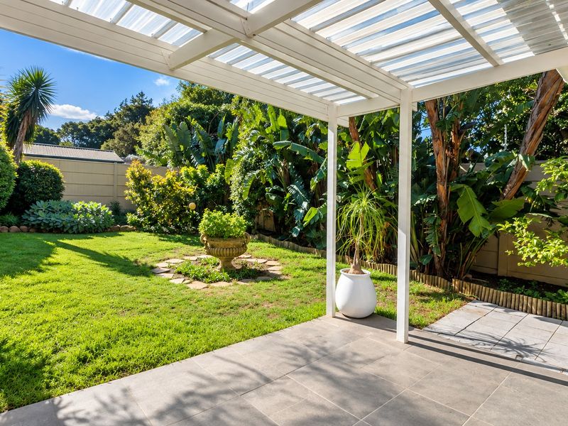 A well-maintained covered patio featuring tiled flooring and a pergola, overlooking a lush, landscaped garden with established greenery and a boundary wall.