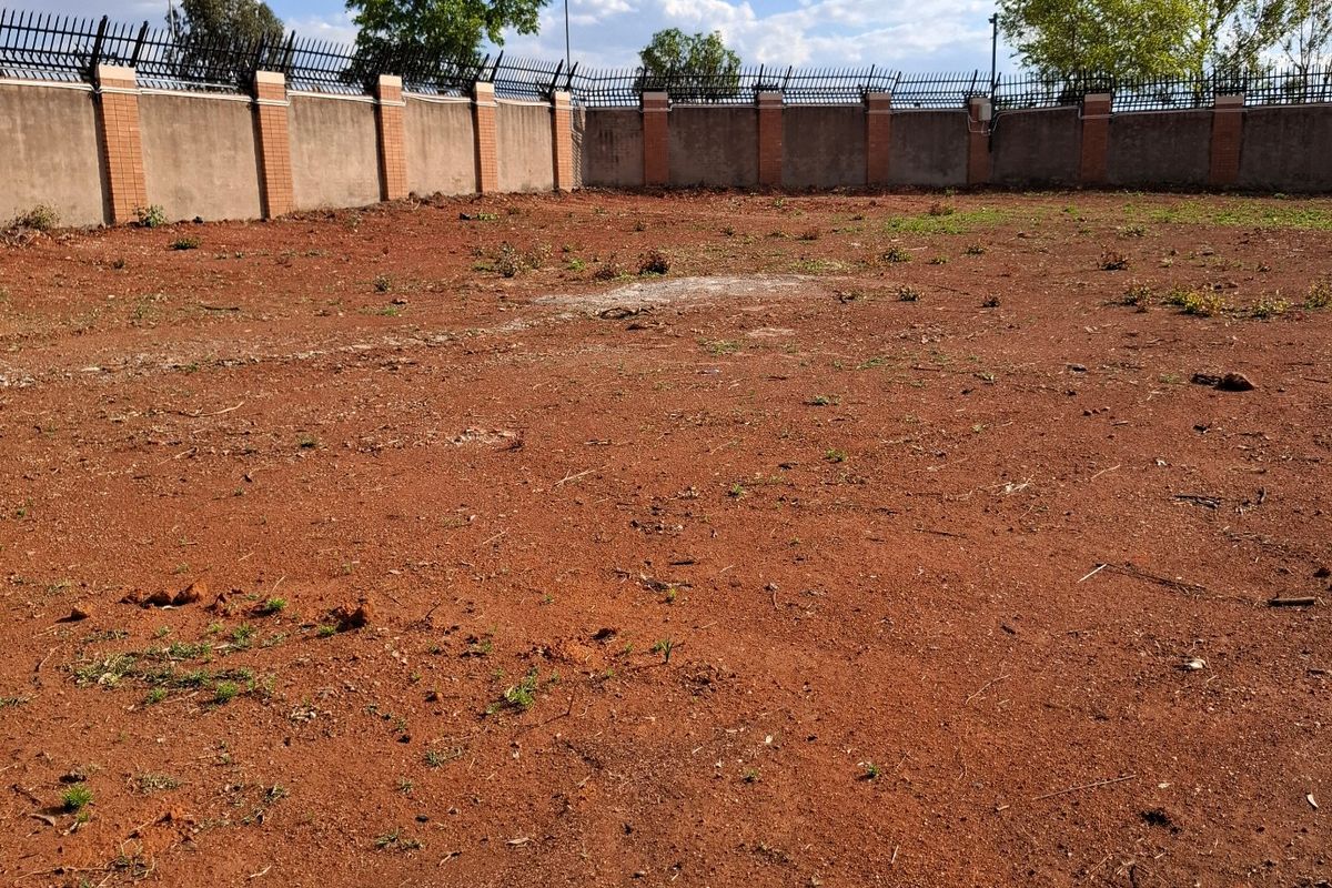 A vacant residential plot featuring a cleared earth surface enclosed by a perimeter wall with integrated brick pillars and security spikes.