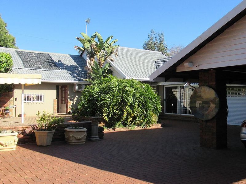A spacious rear exterior area featuring a brick-paved driveway, a covered patio space, and a well-maintained garden setting with solar water heating panels.