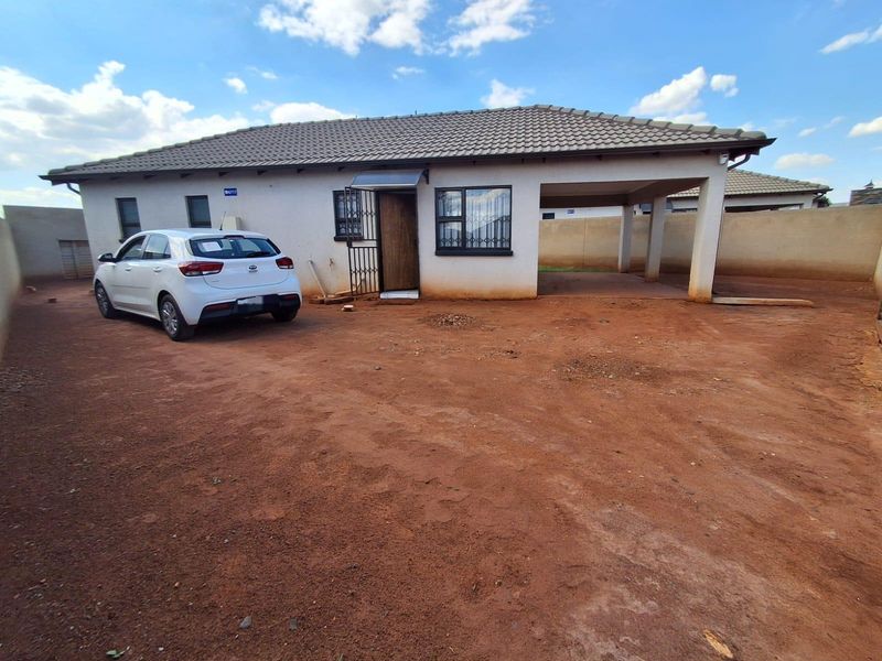 Front view of a single-story house featuring a tiled roof, covered carport, and security gate, set on an unpaved plot.