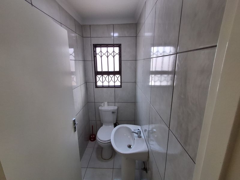 A compact, fully tiled bathroom featuring a pedestal basin and toilet, finished with neutral wall tiles and secure window burglar bars.