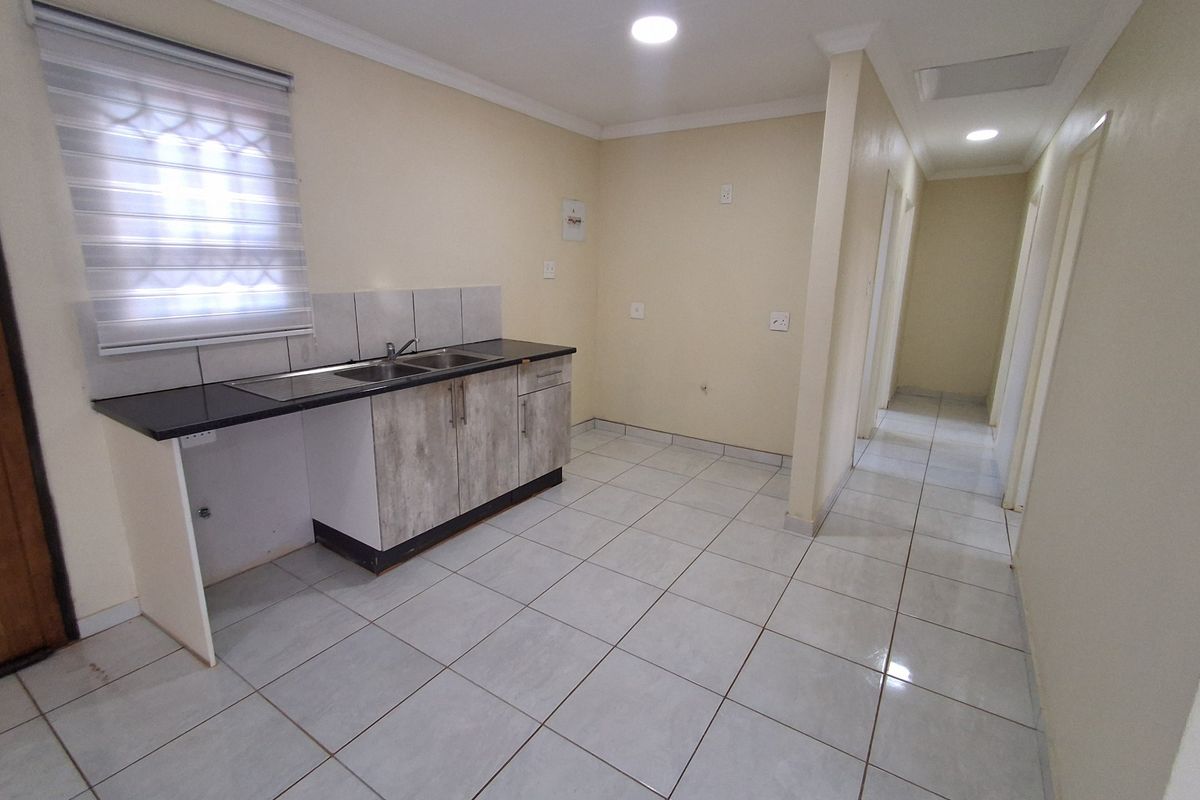 A functional, empty kitchen area featuring tiled flooring, built-in cabinetry with a dark countertop, and a stainless steel sink, leading into a hallway.