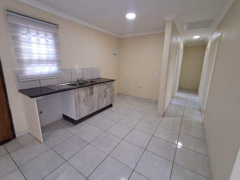 A functional, empty kitchen area featuring tiled flooring, built-in cabinetry with a dark countertop, and a stainless steel sink, leading into a hallway.