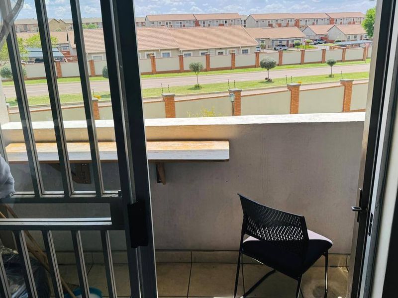 A compact balcony featuring tiled flooring, a security gate, and a wall-mounted shelf, offering an outdoor space with views of the surrounding residential complex.