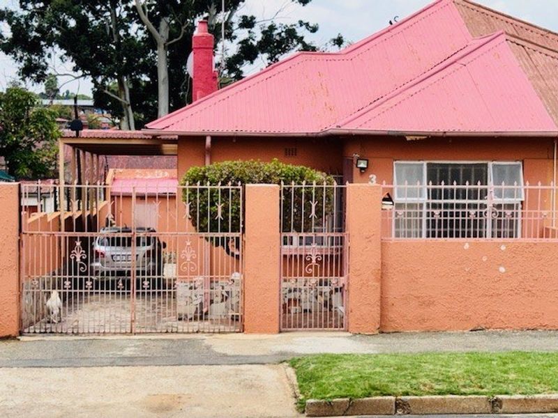 Front elevation of a residential house featuring a corrugated metal roof, brick chimney, and secure wrought iron perimeter fencing with an integrated carport.