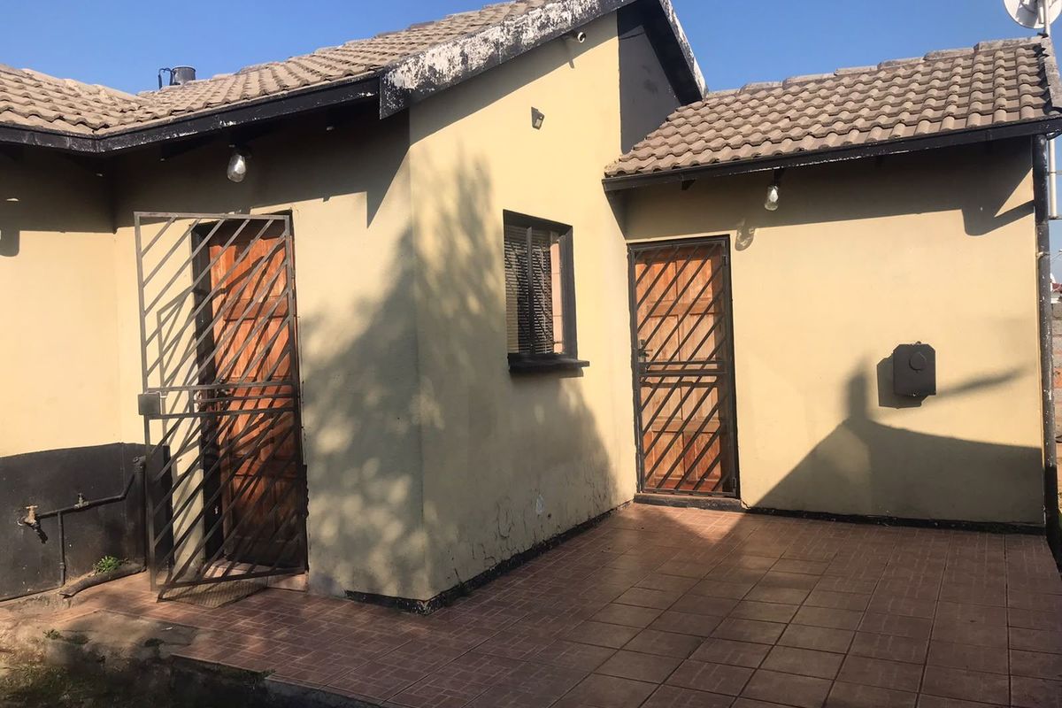 Rear exterior view of the property featuring a tiled patio area, security gates, and wooden doors with visible wear to the roof fascia and walls.