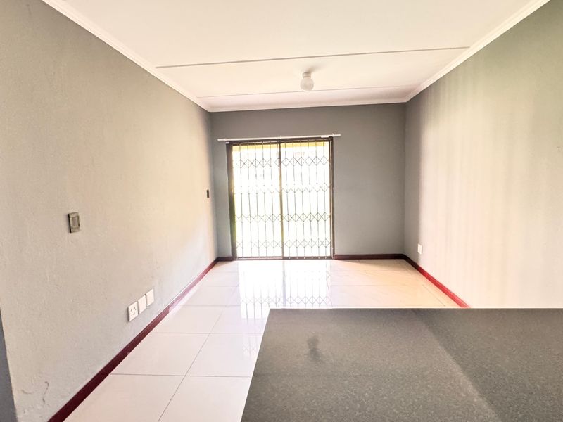 An empty open-plan living area featuring tiled flooring, a kitchen counter, and a sliding door with a security gate leading to the exterior.