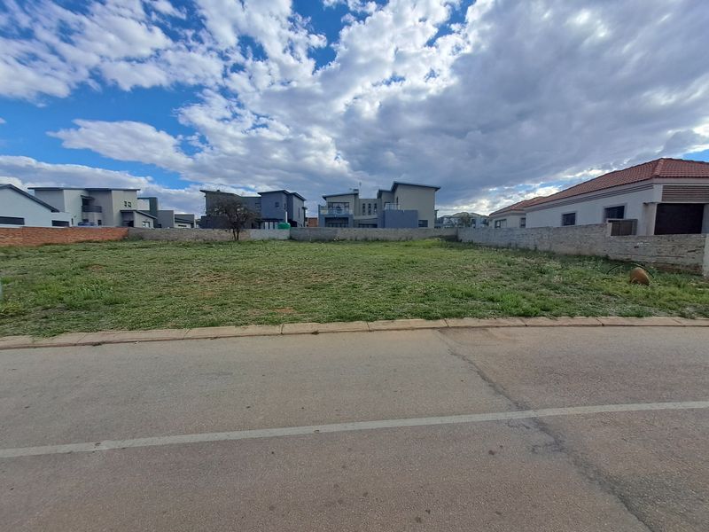 A vacant residential plot of land featuring a boundary wall, situated along a paved street in a developed neighborhood.
