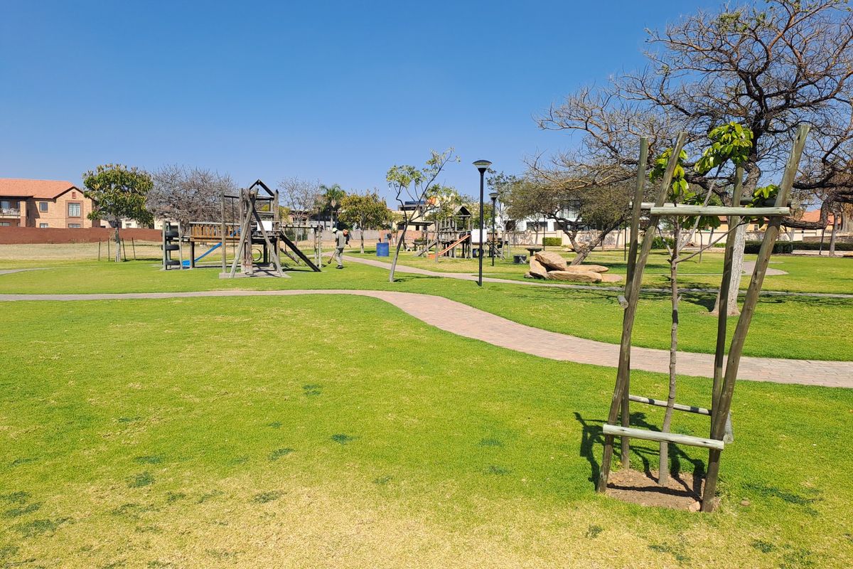 A well-maintained communal garden area featuring paved walkways, established trees, and playground equipment within a residential estate setting.