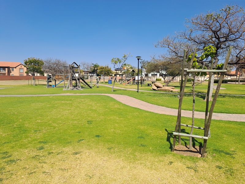 A well-maintained communal garden area featuring paved walkways, established trees, and playground equipment within a residential estate setting.