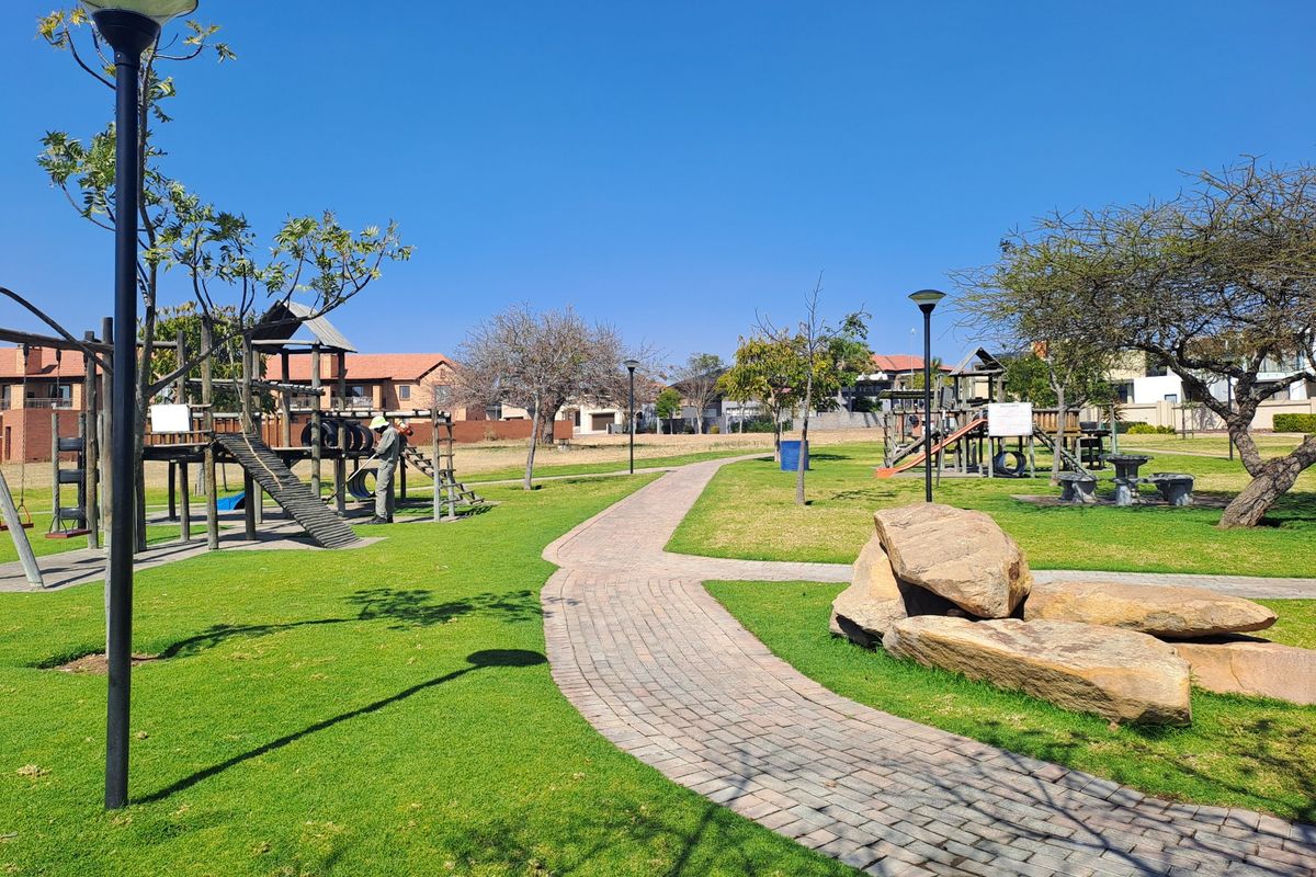 A well-maintained communal garden area featuring a paved walkway, established lawn, decorative rock features, and playground equipment within a residential estate.