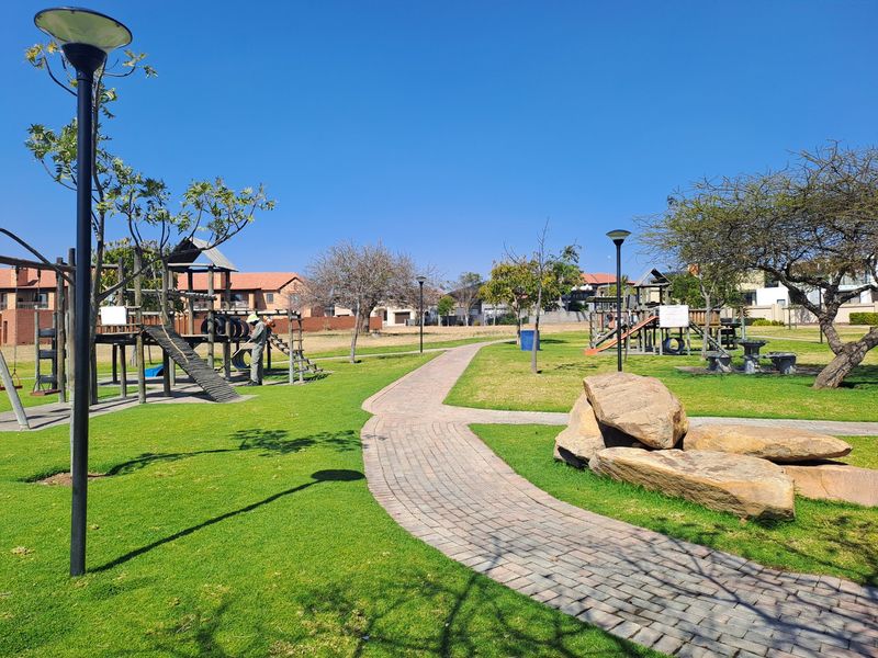 A well-maintained communal garden area featuring a paved walkway, established lawn, decorative rock features, and playground equipment within a residential estate.