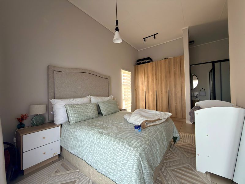 A well-maintained bedroom featuring built-in wooden wardrobes, modern track and pendant lighting, and neutral tiled flooring with an en-suite bathroom access.