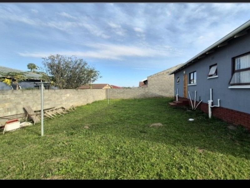 Rear exterior view of the property featuring a large grassed yard, perimeter boundary wall, and exposed external plumbing along the side elevation.