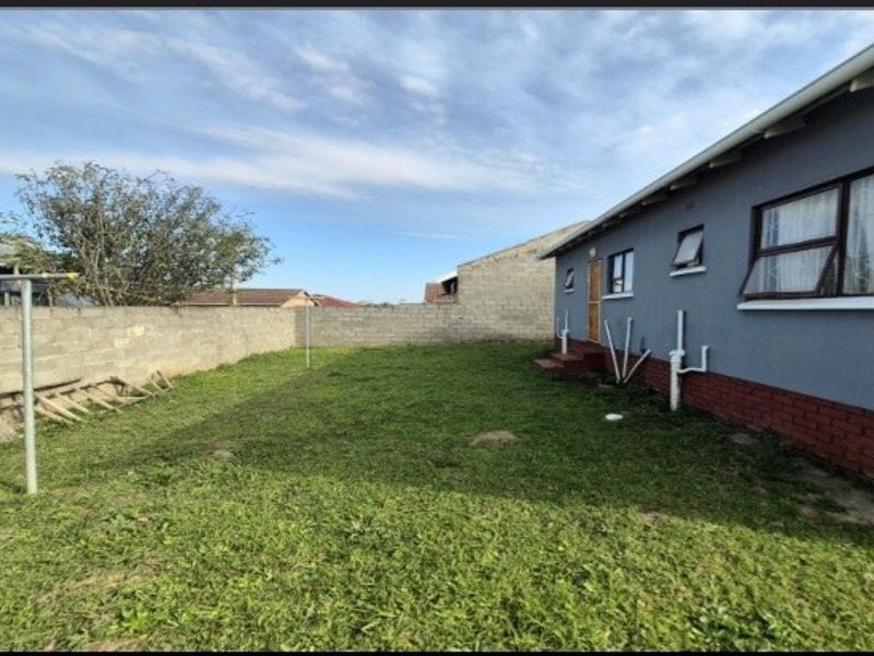 A rear exterior view of the property featuring a grassed backyard, brick boundary wall, and the side elevation of the house with external plumbing.