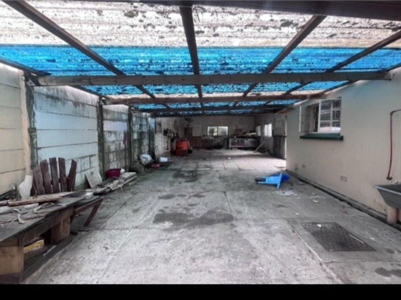 A large, covered outdoor utility or patio area featuring concrete flooring, a utility sink, and a corrugated roof structure in need of maintenance.