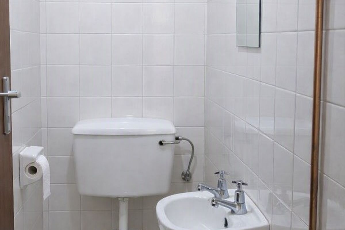 A compact, functional bathroom featuring a white toilet, wall-mounted basin, and fully tiled walls with exposed copper piping.