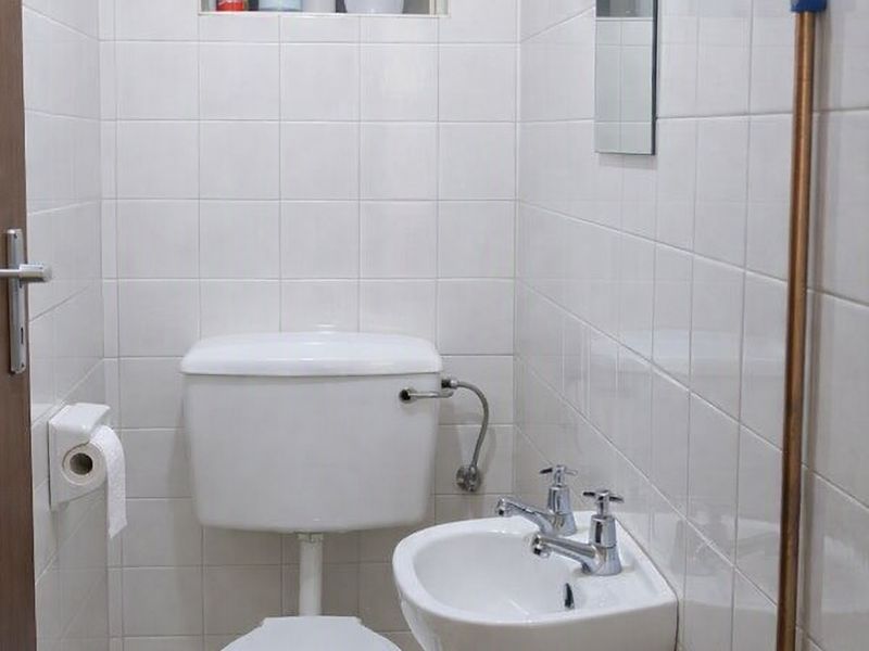 A compact, functional bathroom featuring a white toilet, wall-mounted basin, and fully tiled walls with exposed copper piping.