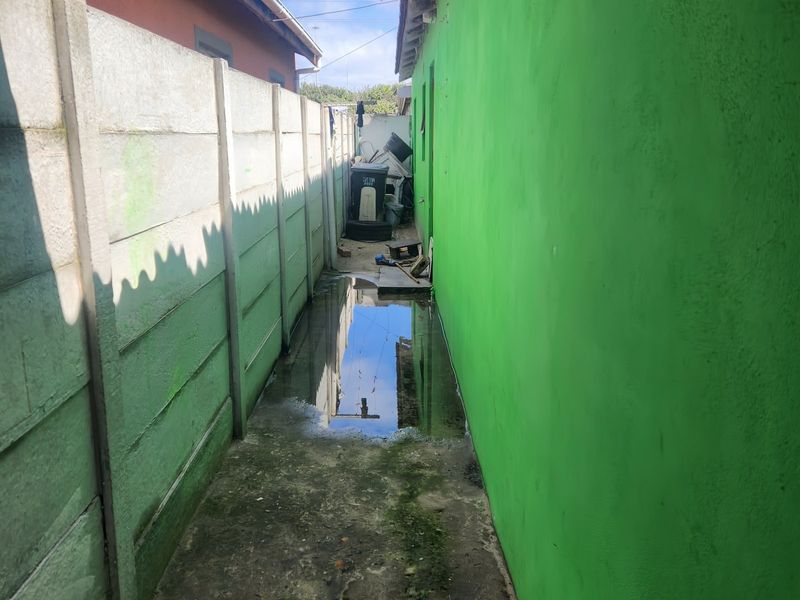 A narrow side passage between the house and a precast concrete boundary wall, showing significant standing water and maintenance issues.