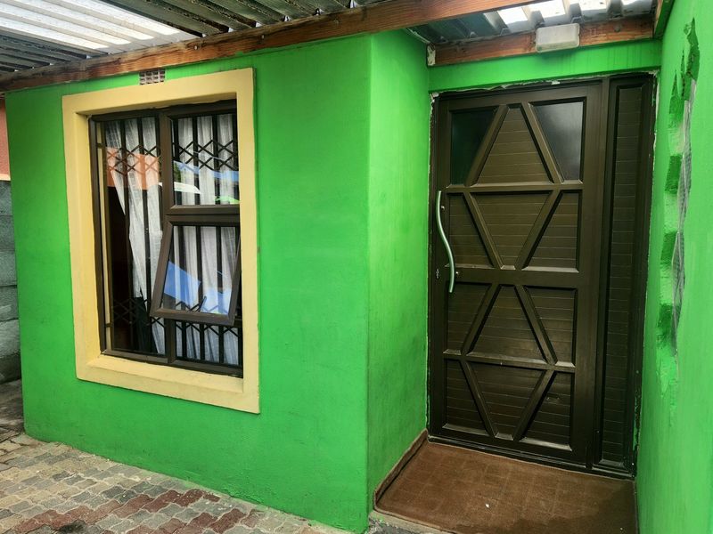 A covered outdoor patio area featuring a security door, window with security bars, and paved flooring, requiring some maintenance to the exterior wall finish.