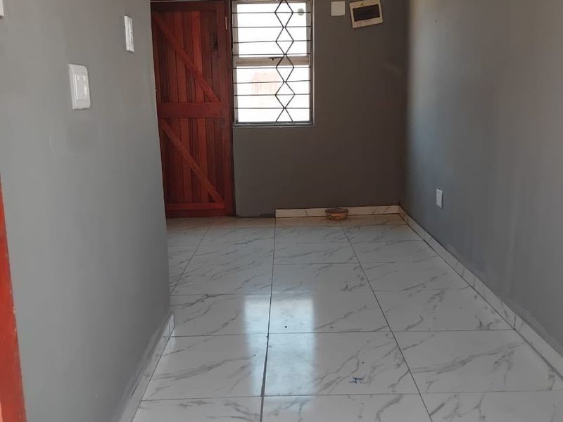 An empty living area featuring marble-look tiled flooring, grey painted walls, and a wooden entrance door with security bars.